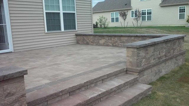 Stone patio with steps leading to a backyard near a beige house and retaining wall.