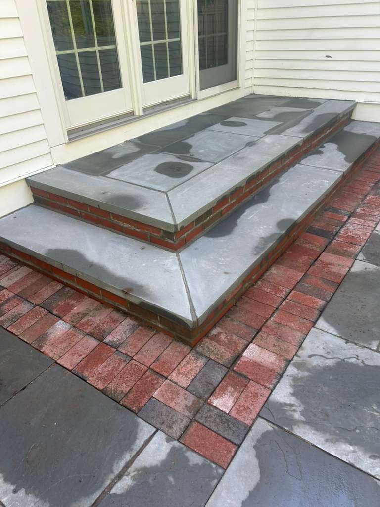 Concrete porch steps and landing with gray slabs and red brick border beside white house siding