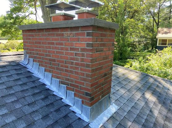 Brick chimney on a gray shingled roof with metal flashing and tree-lined background