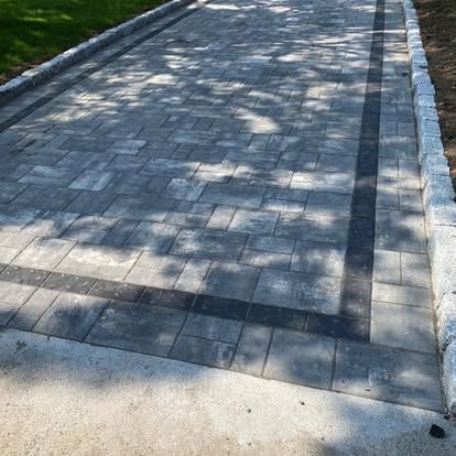 Stone-paved walkway with a dark drainage strip, bordered by grass and curb in dappled sunlight.