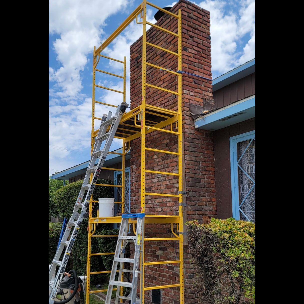 Yellow construction scaffold and ladders beside a brick chimney on a house exterior
