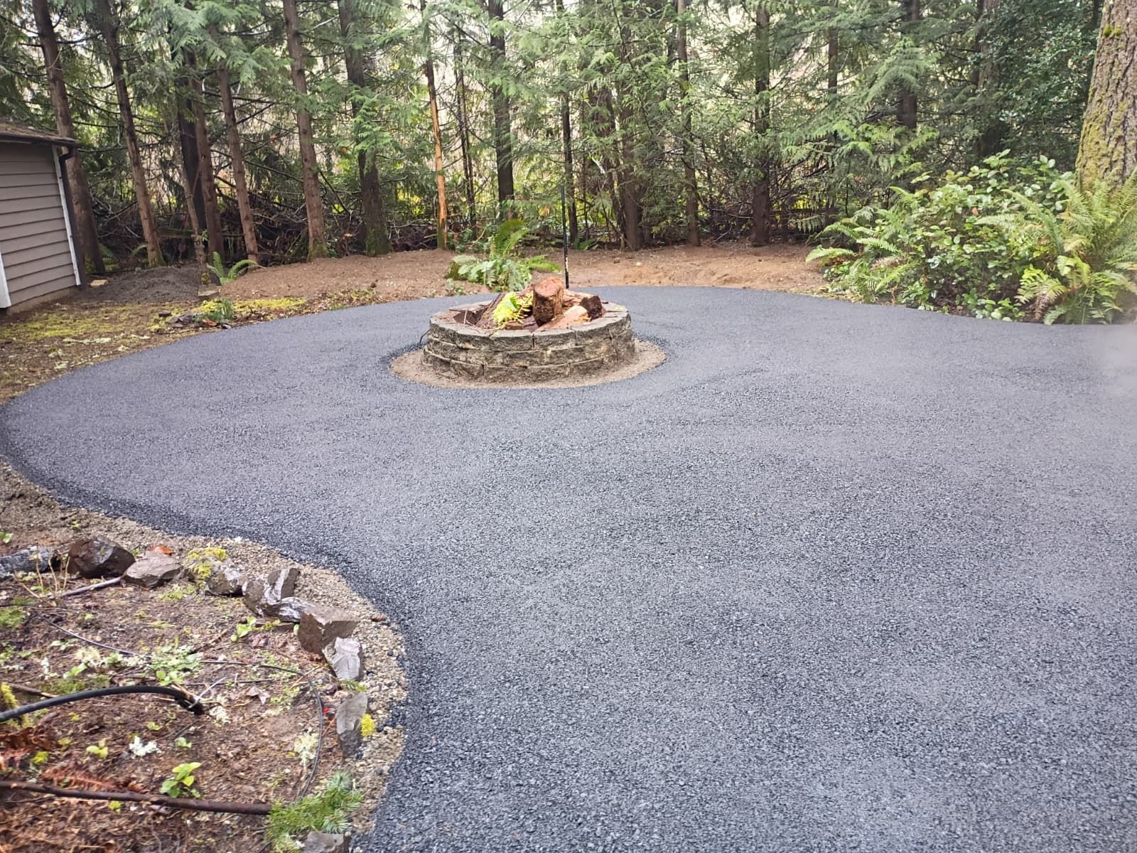 Freshly paved gray circular driveway around a small stone planter in a wooded yard