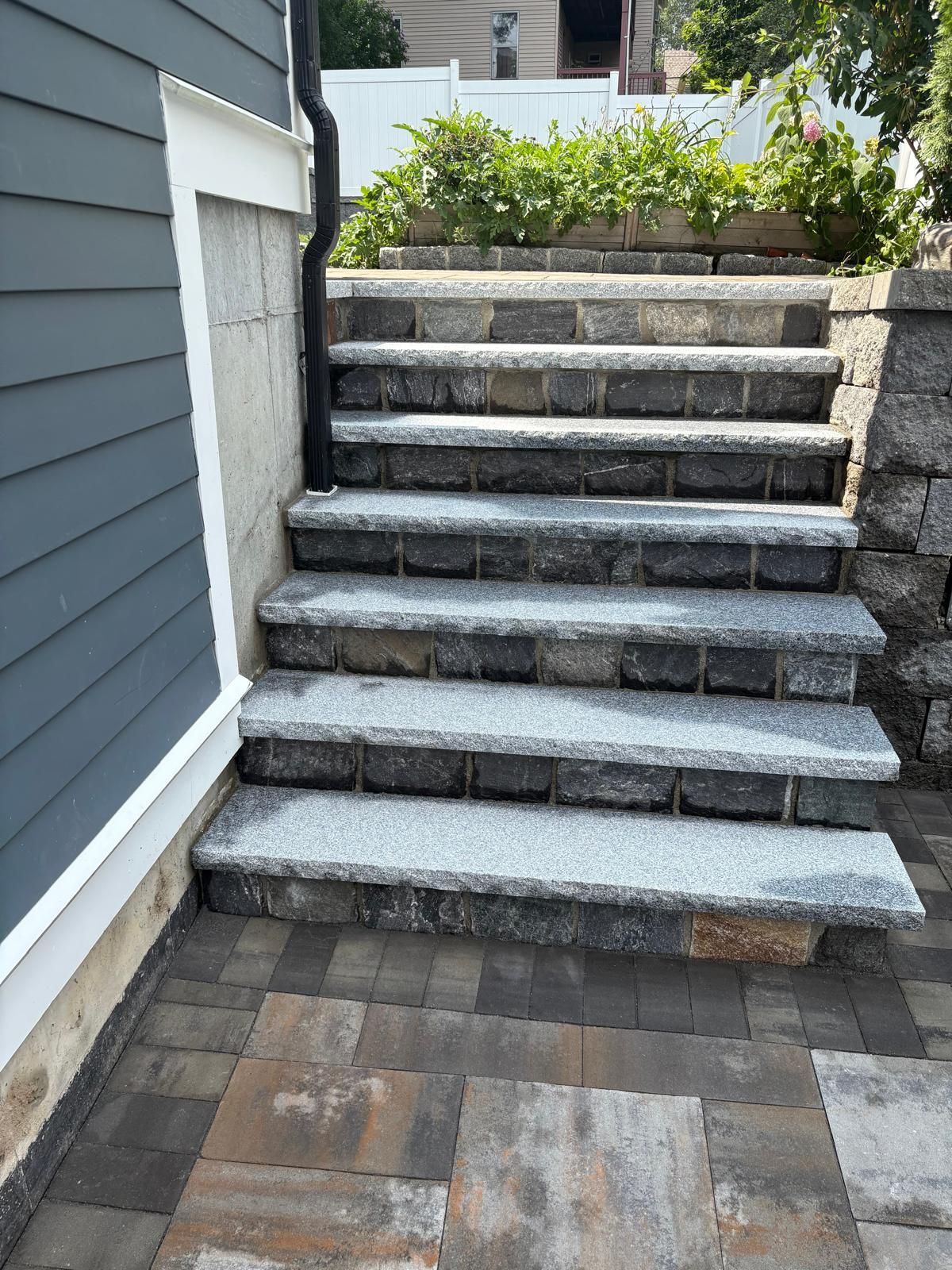 Stone steps leading up to a raised patio beside a gray house, with paver flooring below.