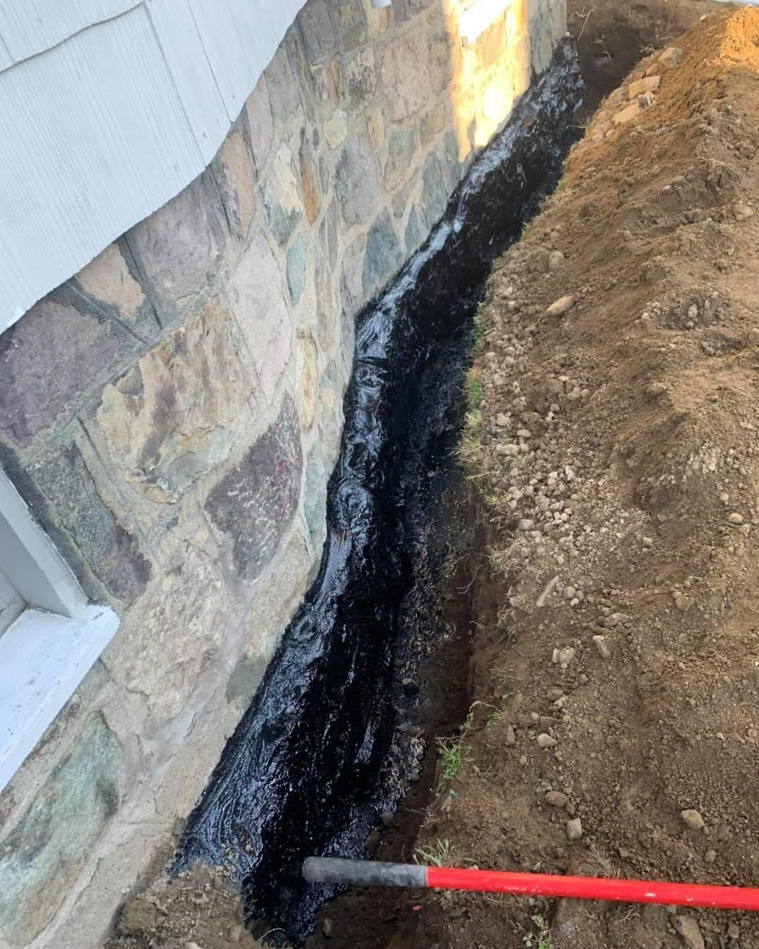 Black waterproofing strip along a stone foundation beside a trench, with a red-handled tool.