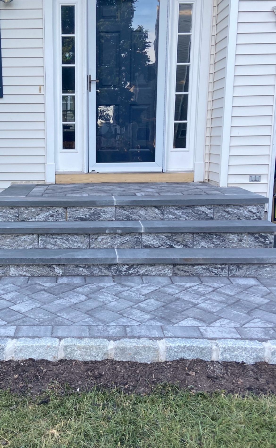 Front entrance with white double doors, stone steps, and a patterned paver walkway leading to a house.