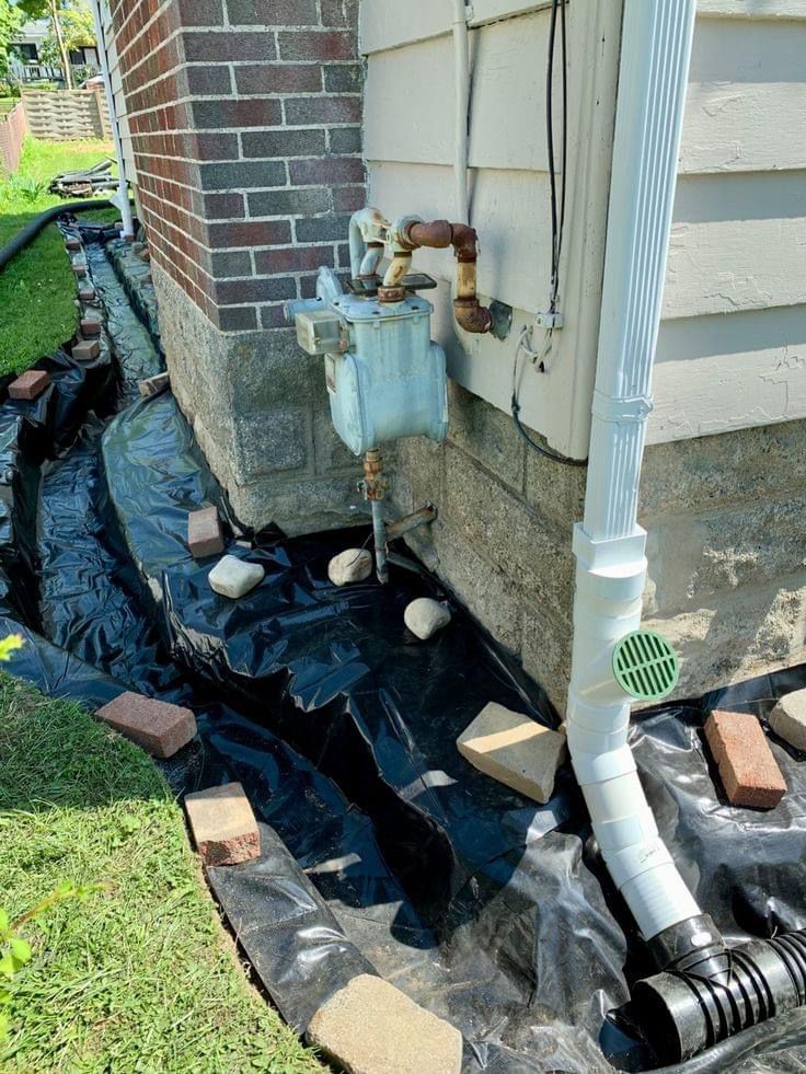 Side of a house with a gas meter, black landscaping fabric, and exposed drain pipe by a brick wall.