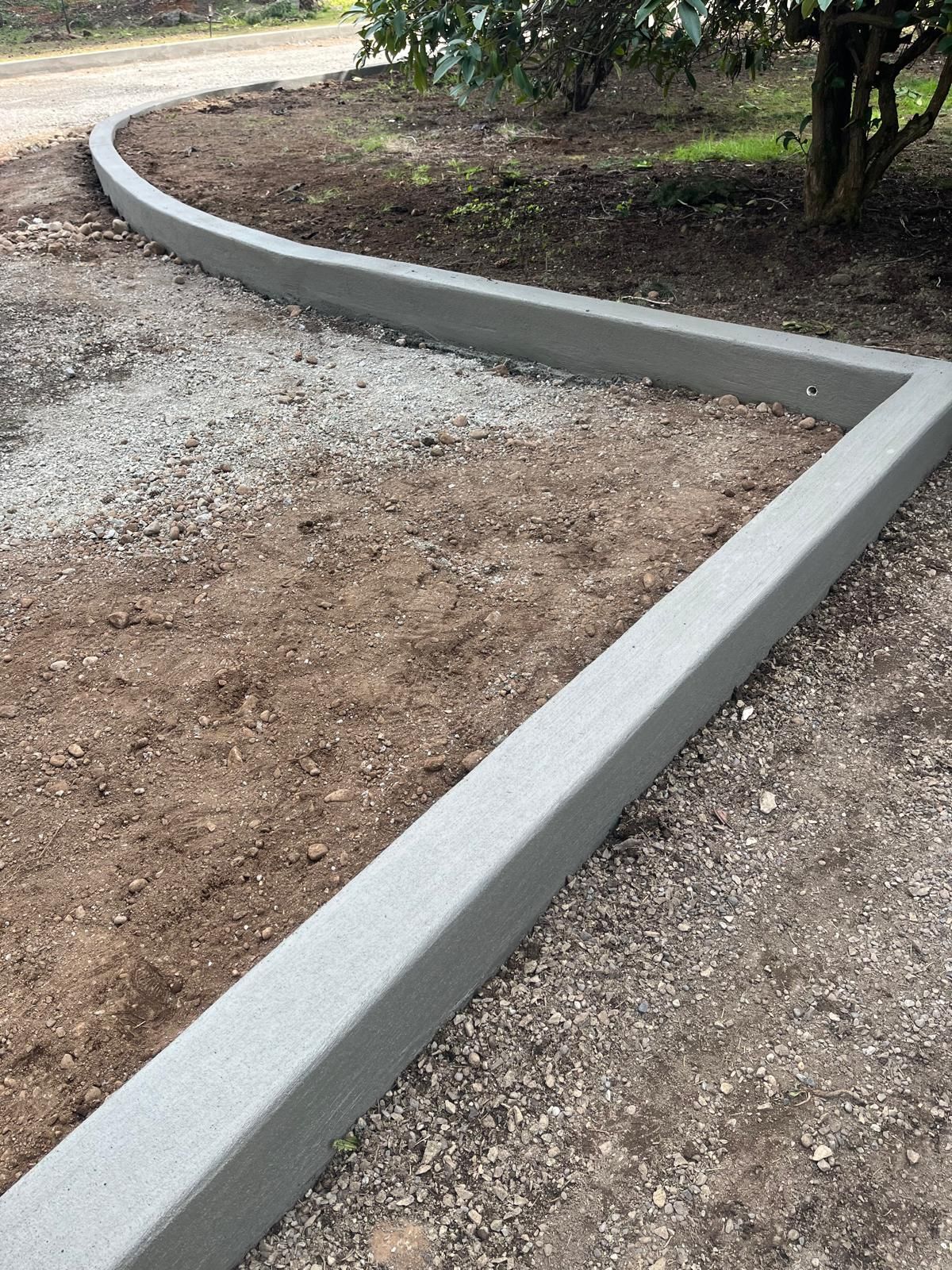 Concrete curb edging along a gravel and dirt path in a garden area, curving around a tree.