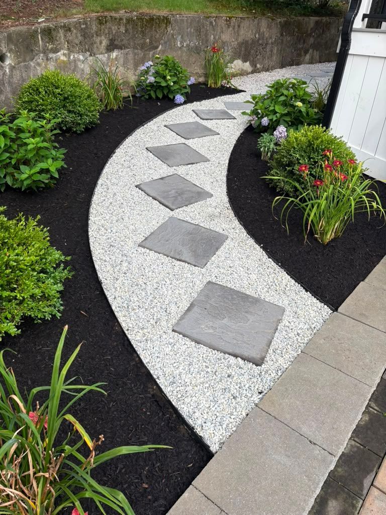 Curved garden path of white gravel and gray stepping stones, bordered by black mulch and green plants.