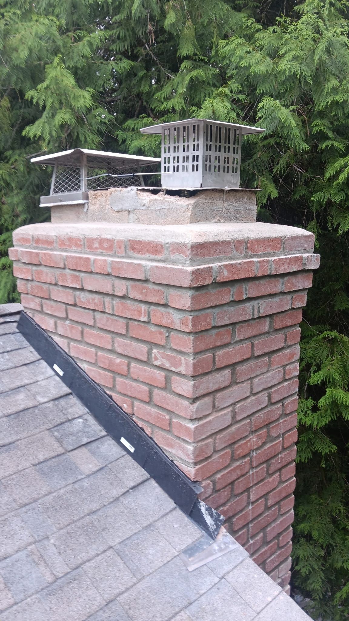 Brick chimney with a metal vent cap on a shingled roof, backed by green trees.