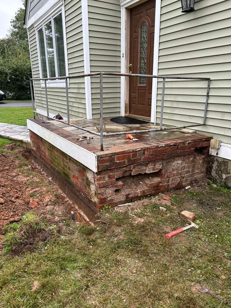 Brick porch with white railing and steps beside a house, damaged and uneven.