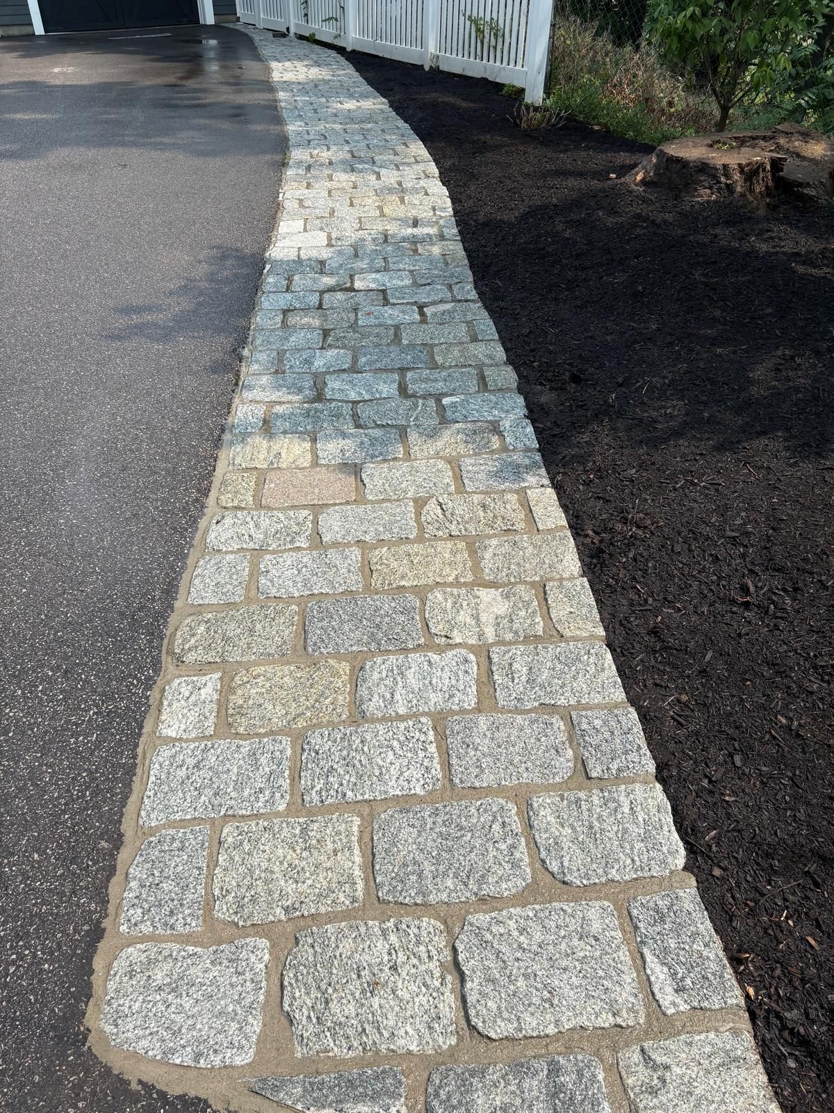 Curved stone walkway bordered by dark mulch and a white fence on one side