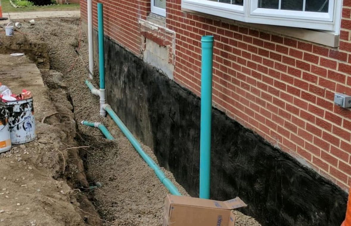 Exterior brick wall with black waterproofing, blue drainage pipes, and a dug trench beside a house