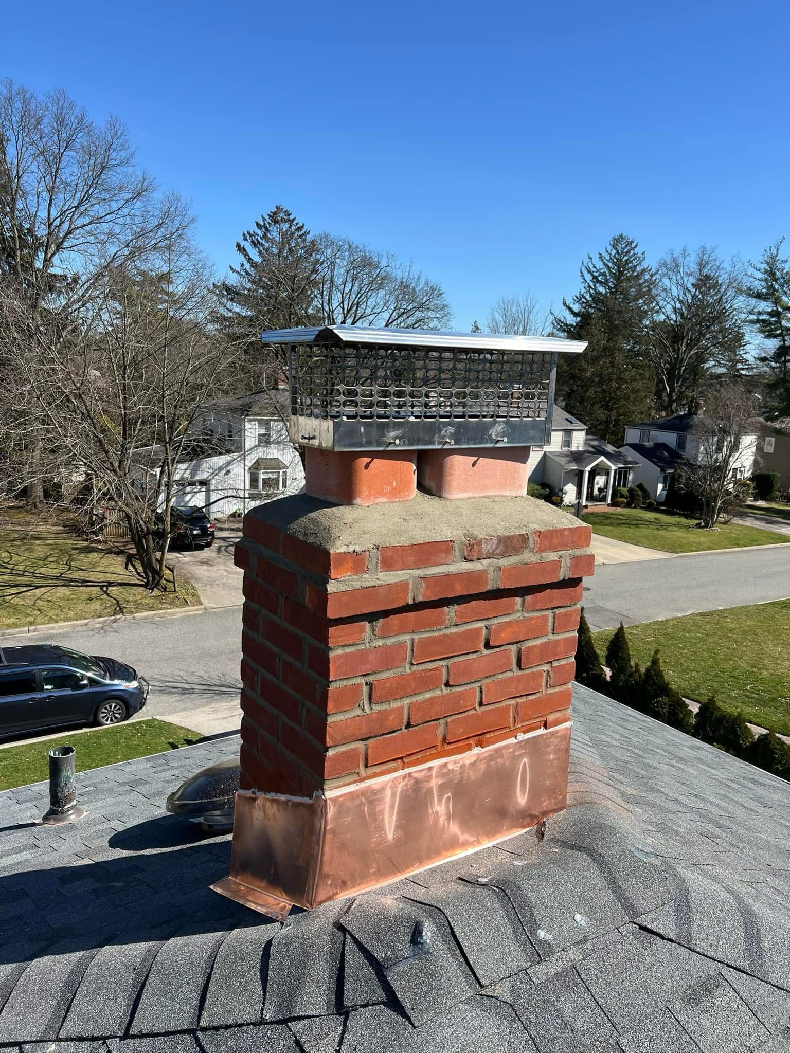 Brick chimney on a shingled roof with houses and trees in the background under a clear blue sky