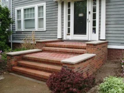 Brick front steps with white trim leading to a gray house entrance, bordered by shrubs and landscaping.