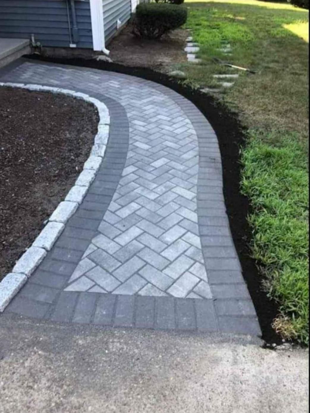 Curved gray paver walkway beside a house, bordered by dark edging and grass.
