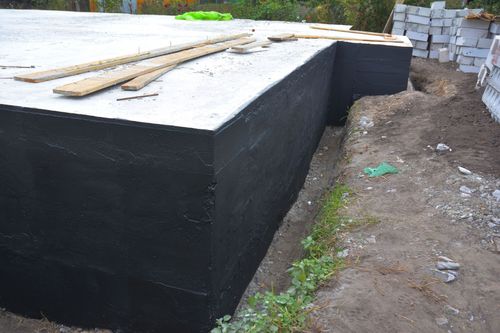 Large black concrete block foundation beside a dirt path, with wooden planks on top and stacked blocks in back