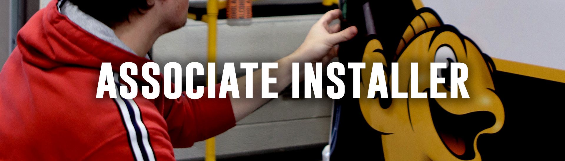 Man applying a decal with a yellow fish logo on a car's rear. He wears a red hoodie and jeans, in a garage.
