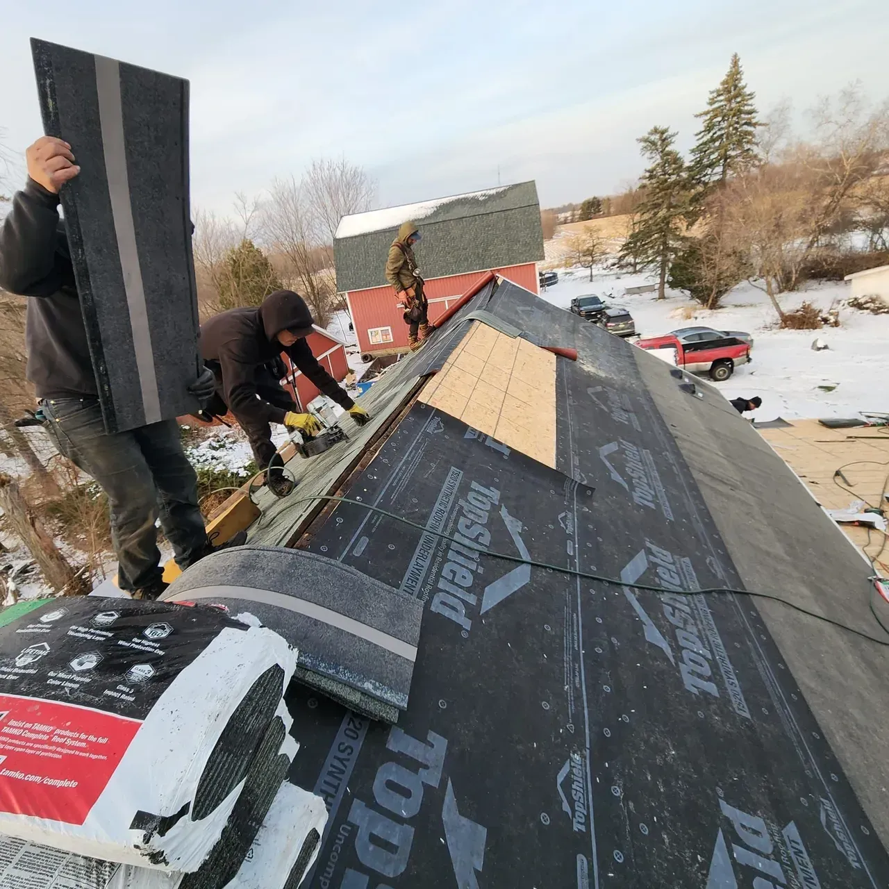 A man is standing on top of a roof holding a piece of top shield shingles
