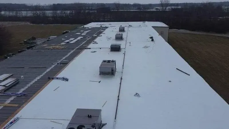 Aerial view of a flat, white roof with HVAC units, some snow, and a surrounding rural landscape.