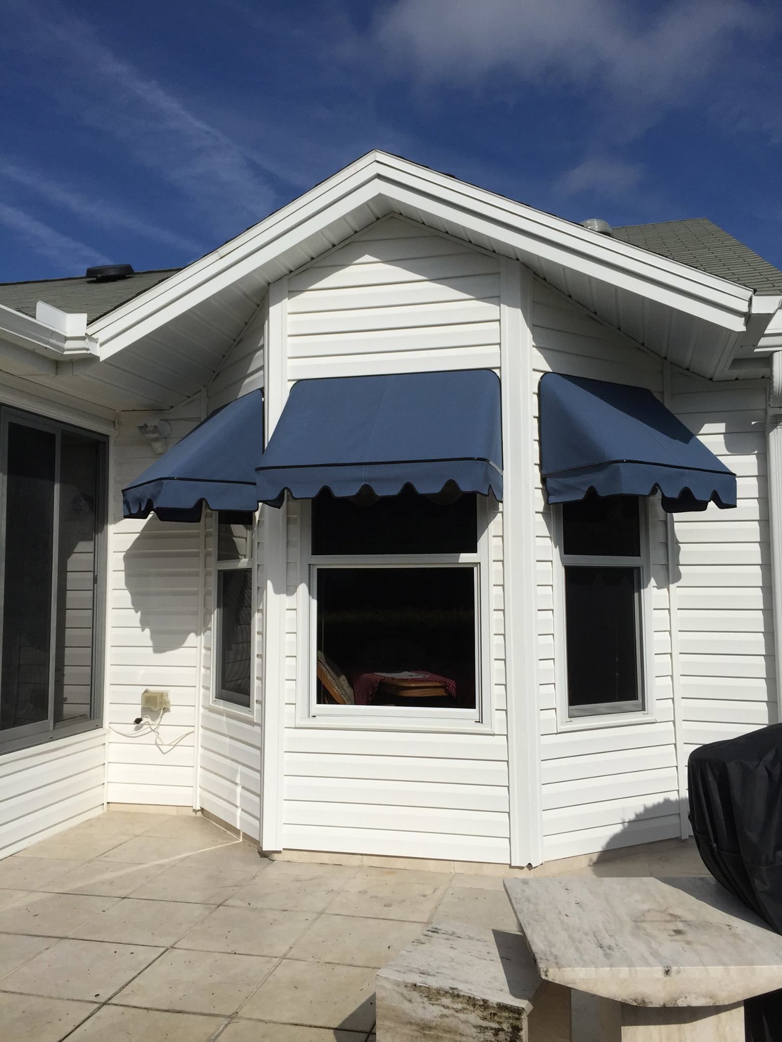 Three blue fabric window awnings with scalloped edges installed on the exterior of a white, paneled bay window house wall.