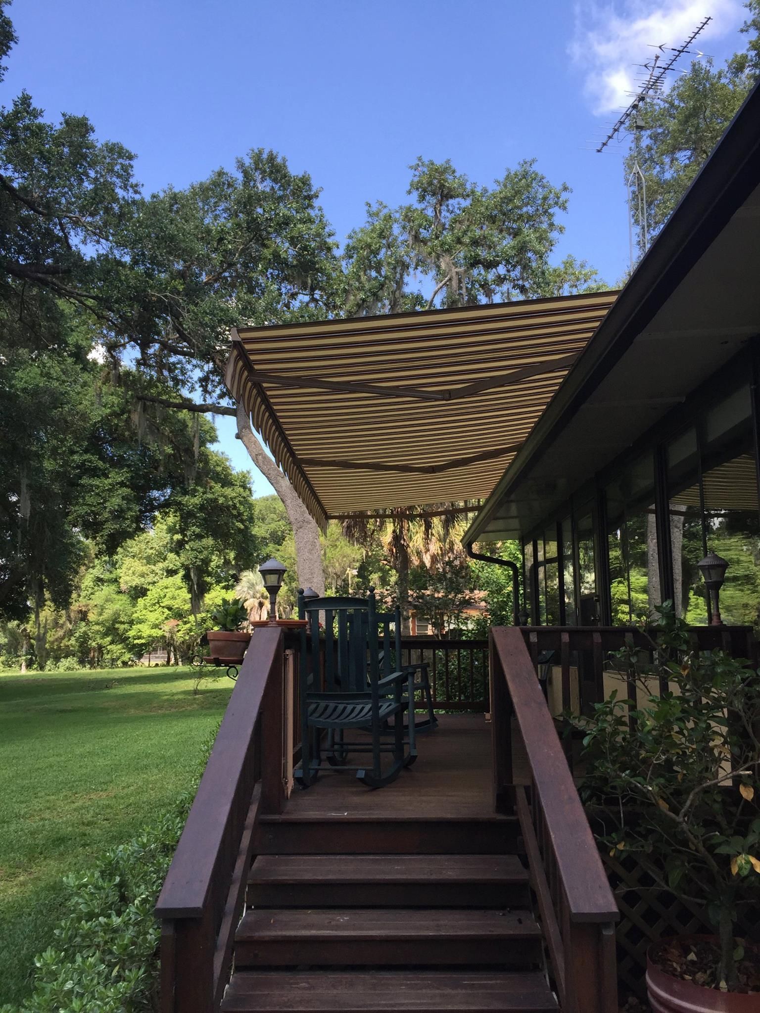 A wooden deck with a rocking chair under a patterned canopy, overlooking a green yard with trees under a blue sky.
