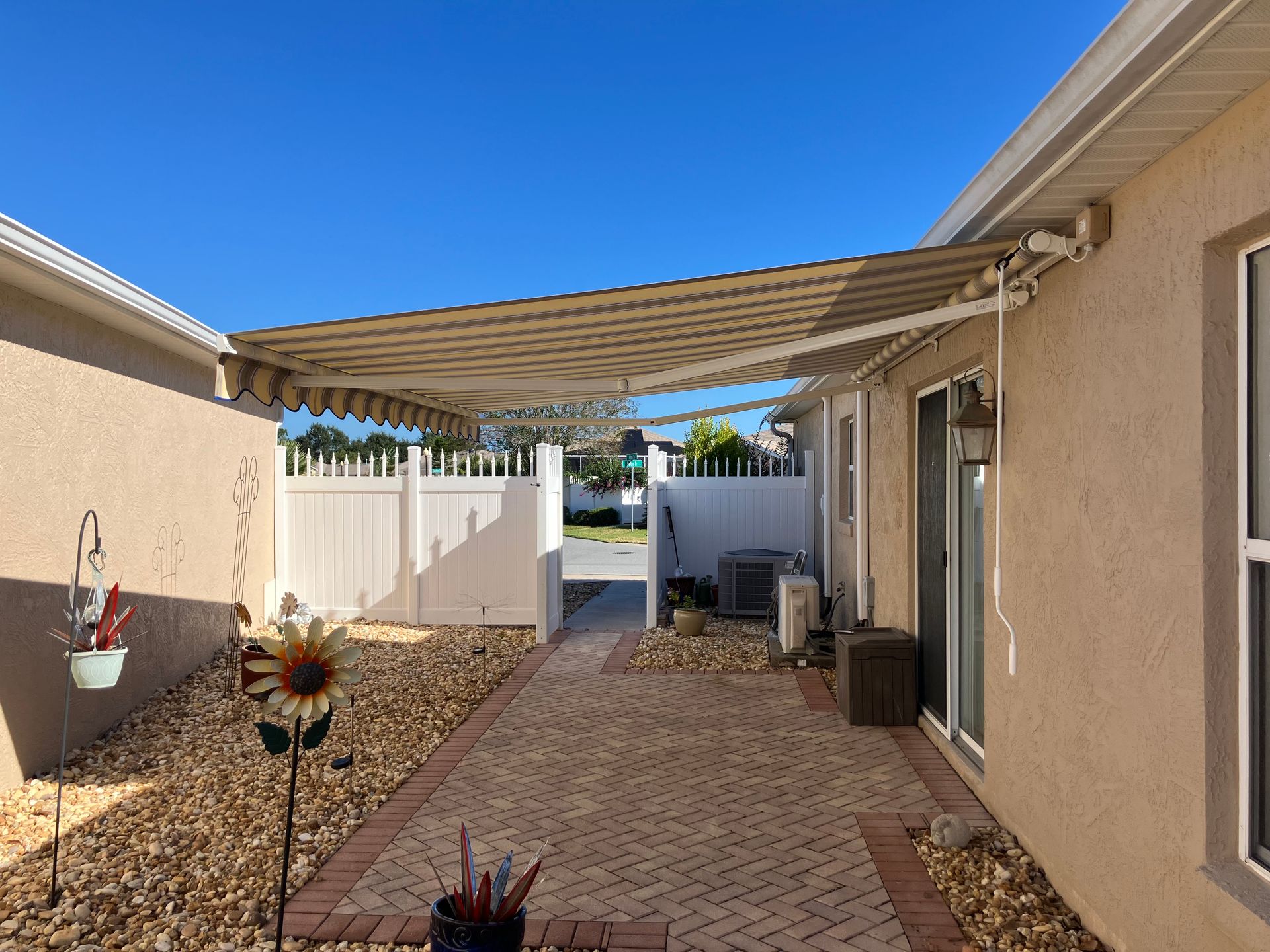 A beige striped retractable awning covers a brick patio between two stucco houses on a bright, sunny day.