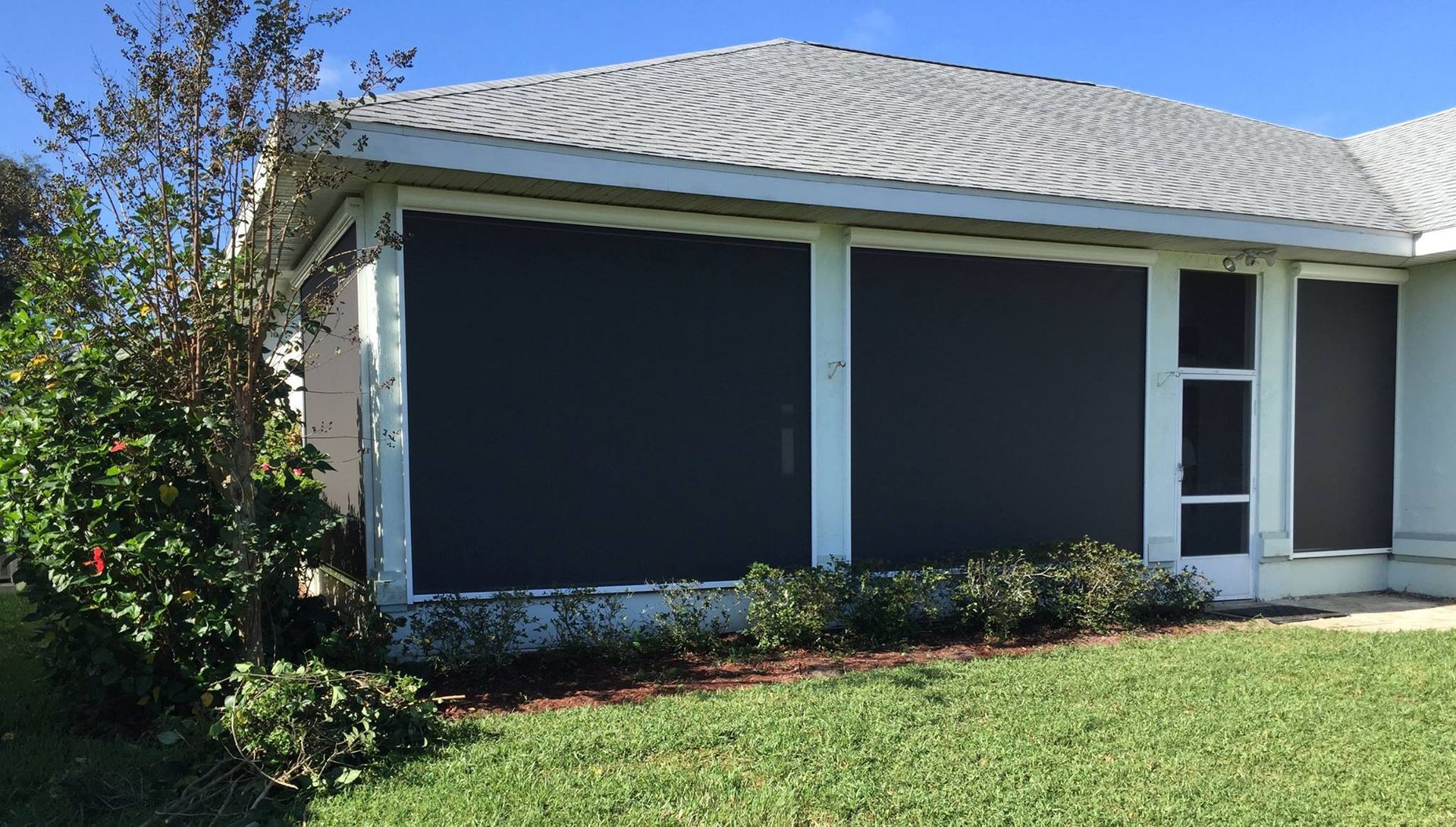 A house exterior featuring a covered patio with dark sun-blocking shades, white columns, and a landscaped lawn.