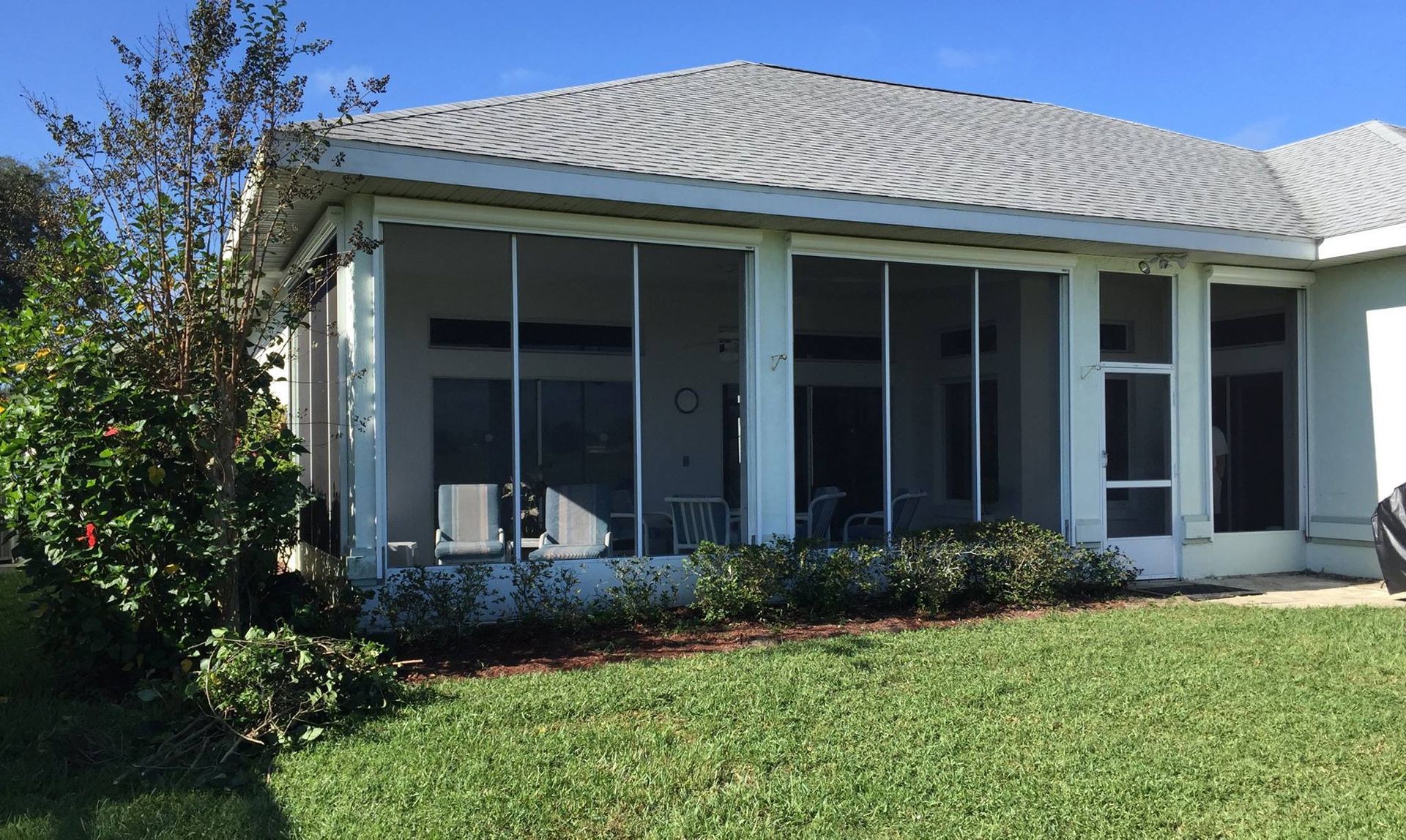 A screened-in patio attached to a light-colored house with a shingled roof, set beside a grassy lawn and a green bush.