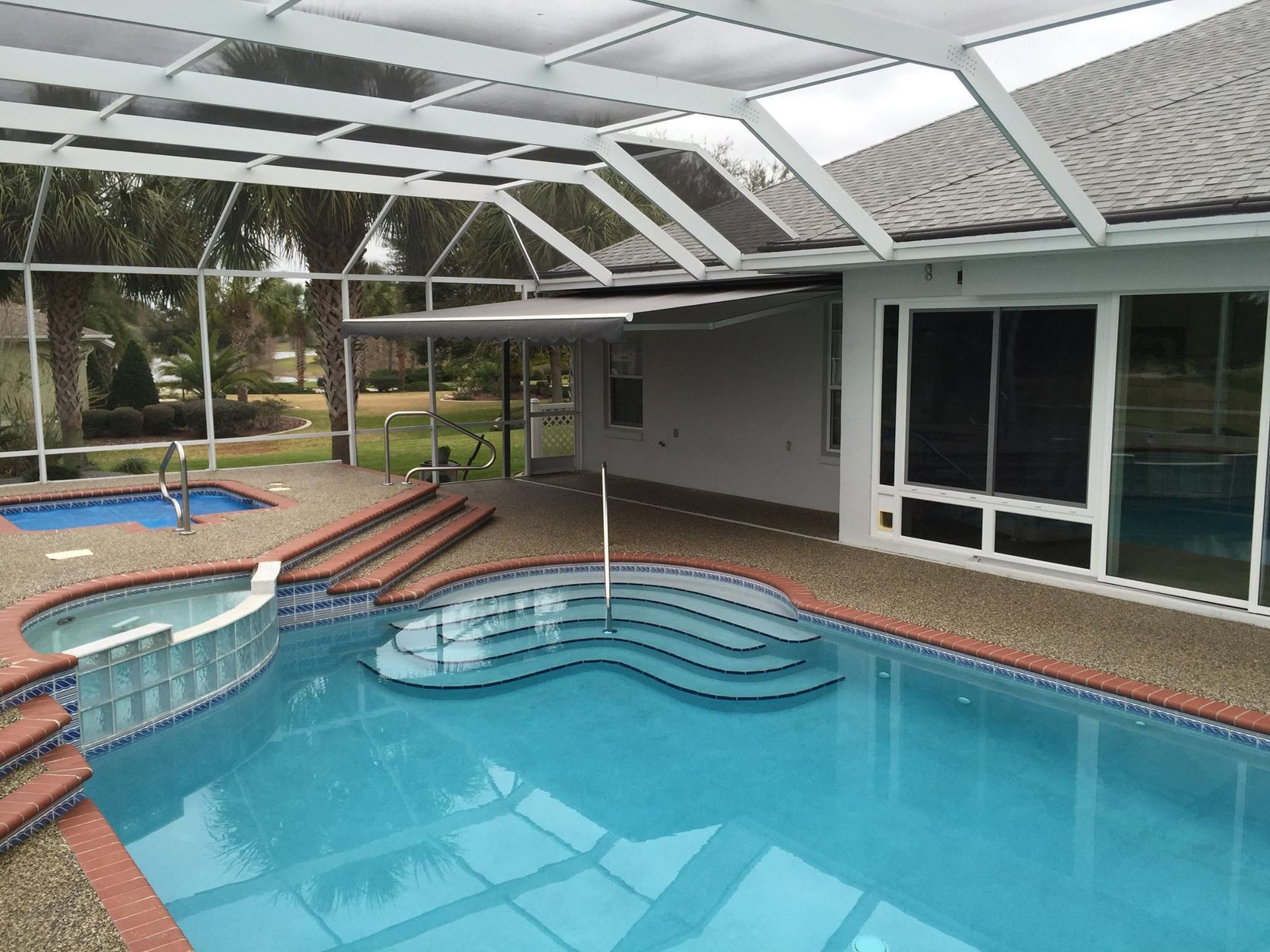 A screened-in patio area featuring a swimming pool with tiered steps, a raised hot tub, and a retractable awning.
