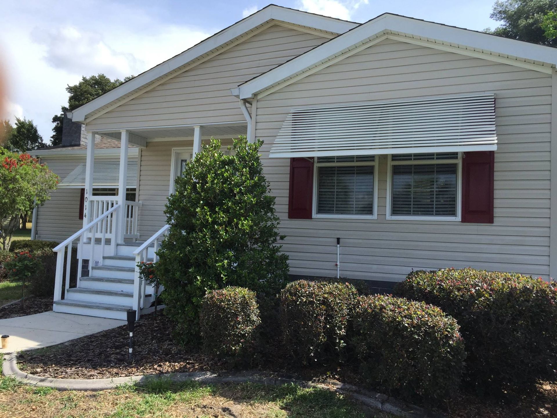 A one-story beige house with a front porch, white railings, a striped window awning, and a red-trimmed window.