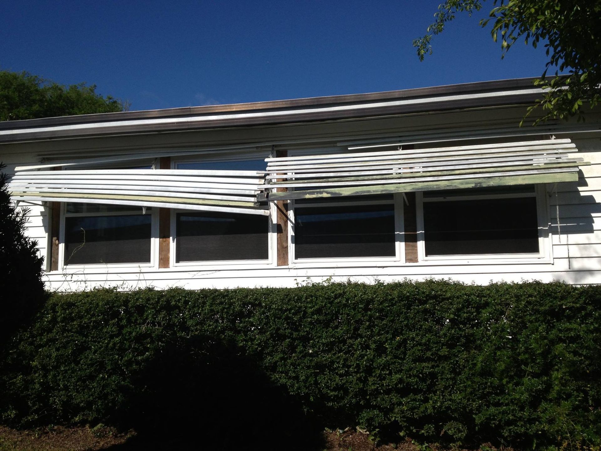A house exterior with white siding and large windows, obscured by a damaged, horizontally sagging metal window awning.