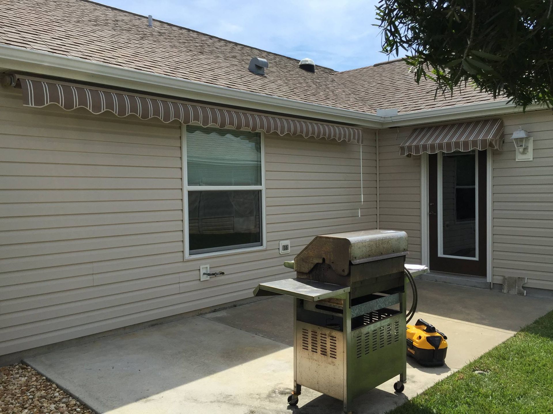 A stainless steel grill sits on a concrete patio in front of a house with beige siding, a window, and retractable awnings.