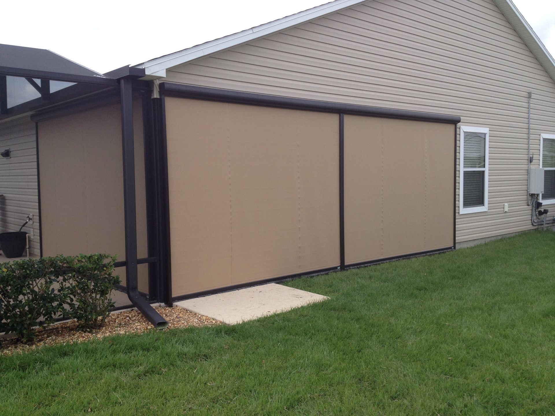 Tan retractable privacy screens installed along the side of a beige, vinyl-sided suburban house near a grassy lawn.