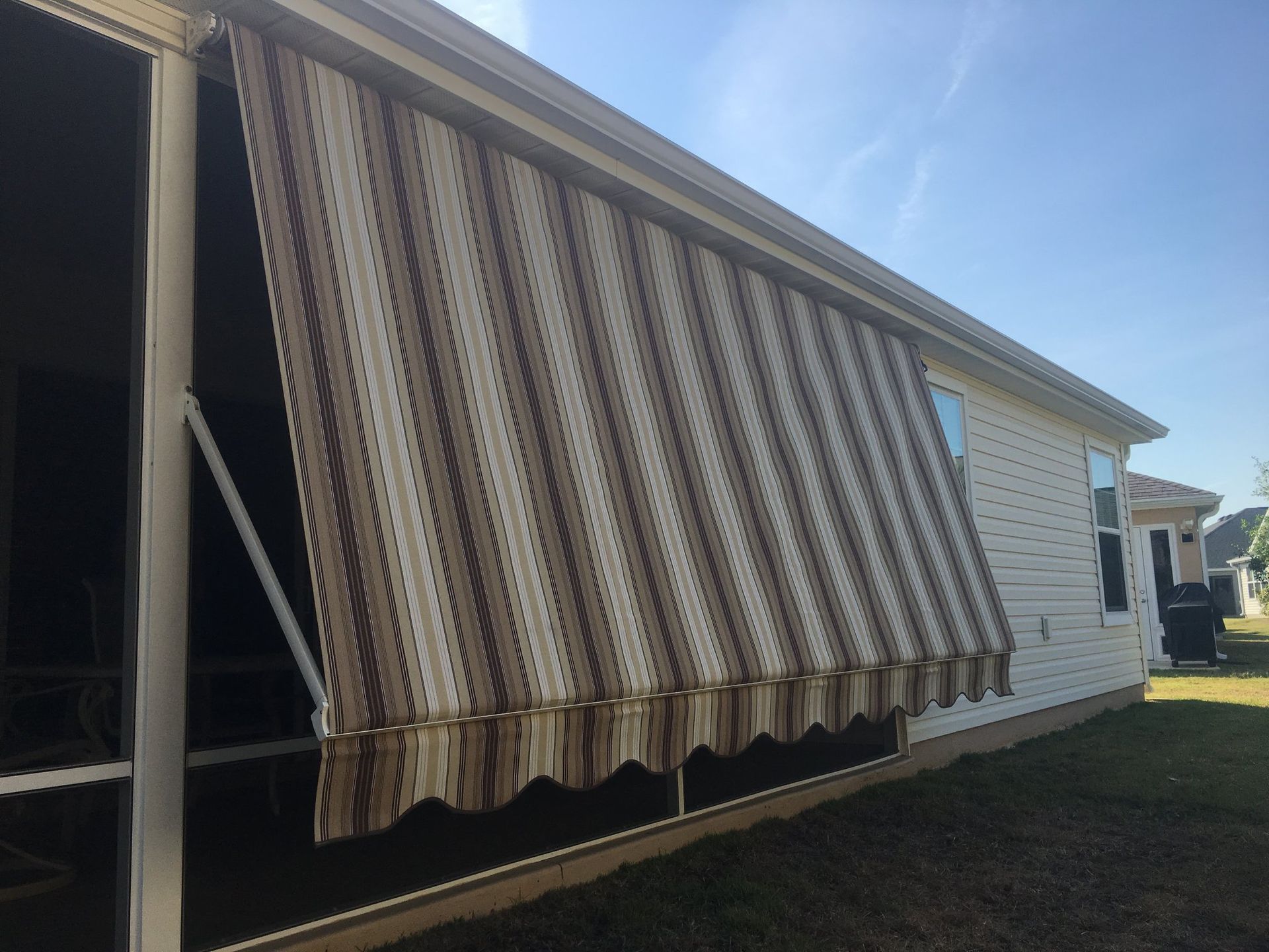 A brown and tan striped retractable awning extended over the side of a white house on a sunny day.