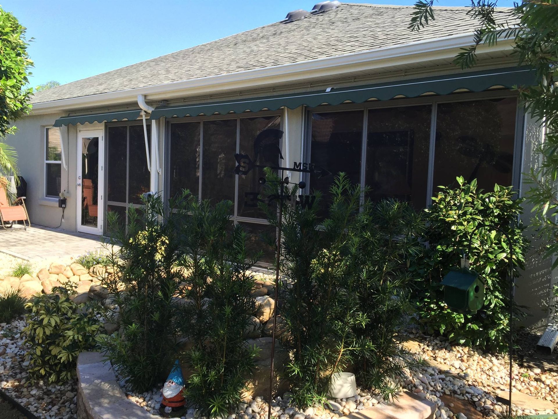 A rear view of a light-colored house featuring a large screened-in porch, surrounded by landscaping and garden ornaments.