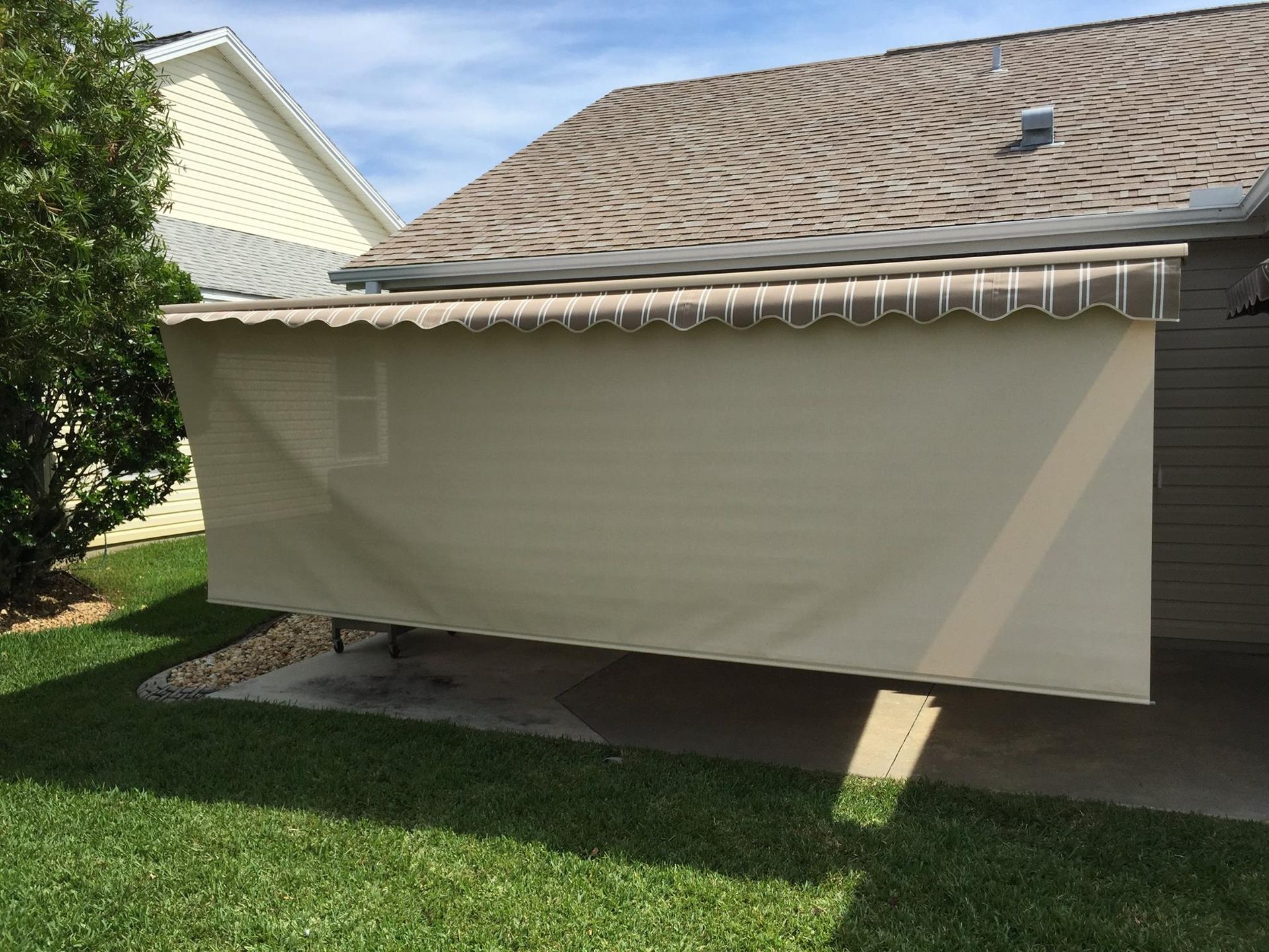 Beige patio retractable awning with a drop-down privacy shade attached to the side of a residential house.