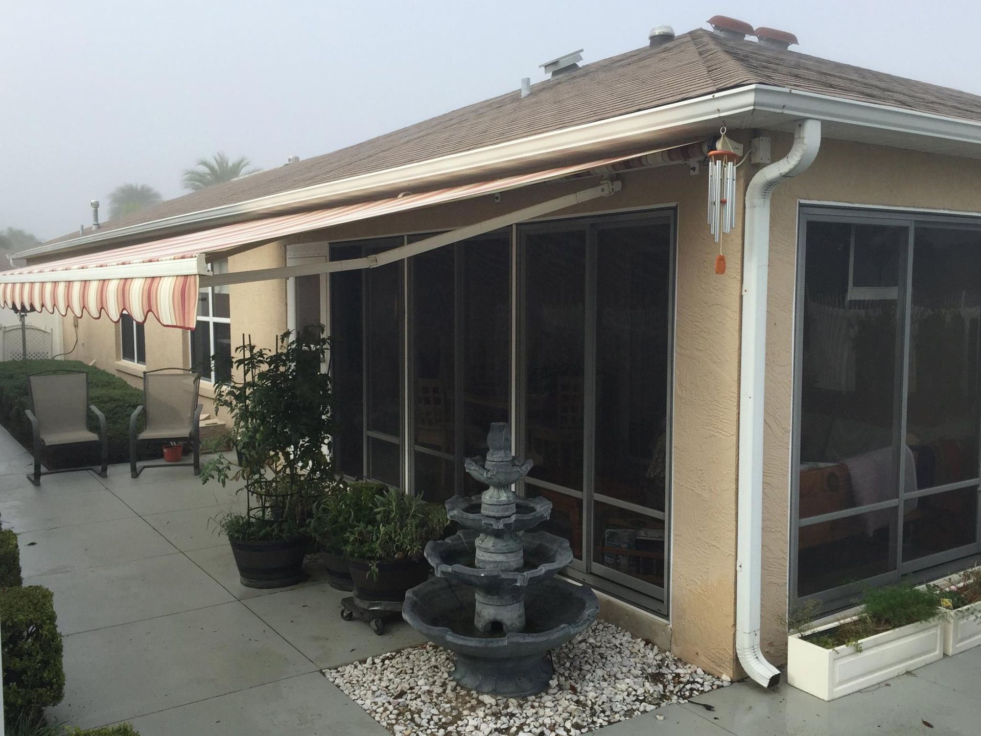 A tan house exterior with a striped retractable awning, patio furniture, a tiered stone fountain, and potted plants.