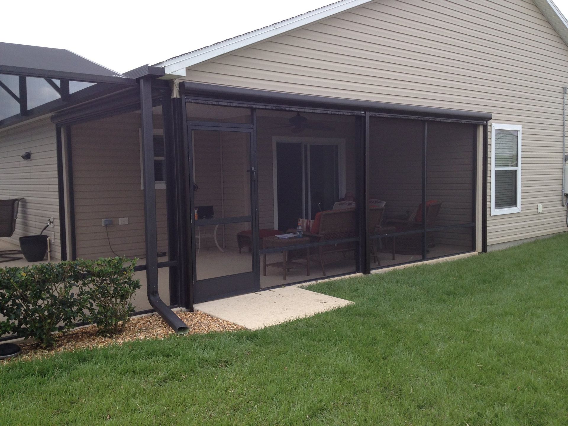 A tan house with a black-framed, screened-in patio enclosure featuring a door, situated on a green lawn.