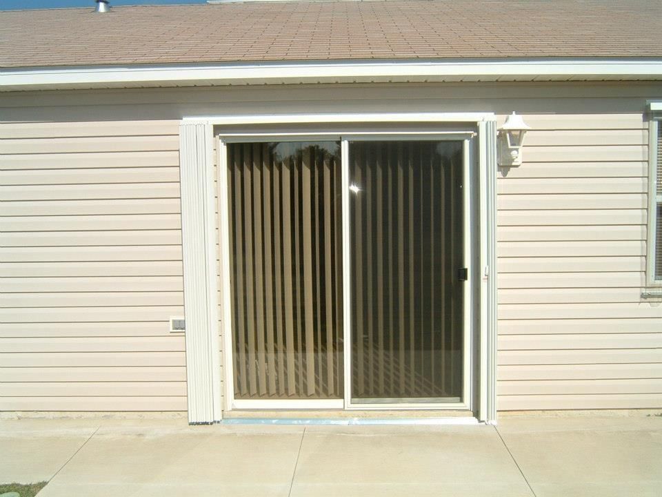A sliding glass door with beige vertical blinds set into the light-colored vinyl-sided exterior of a house.