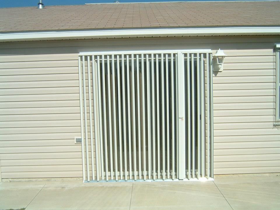 A white metal security gate covering a sliding glass door on the beige siding wall of a house.