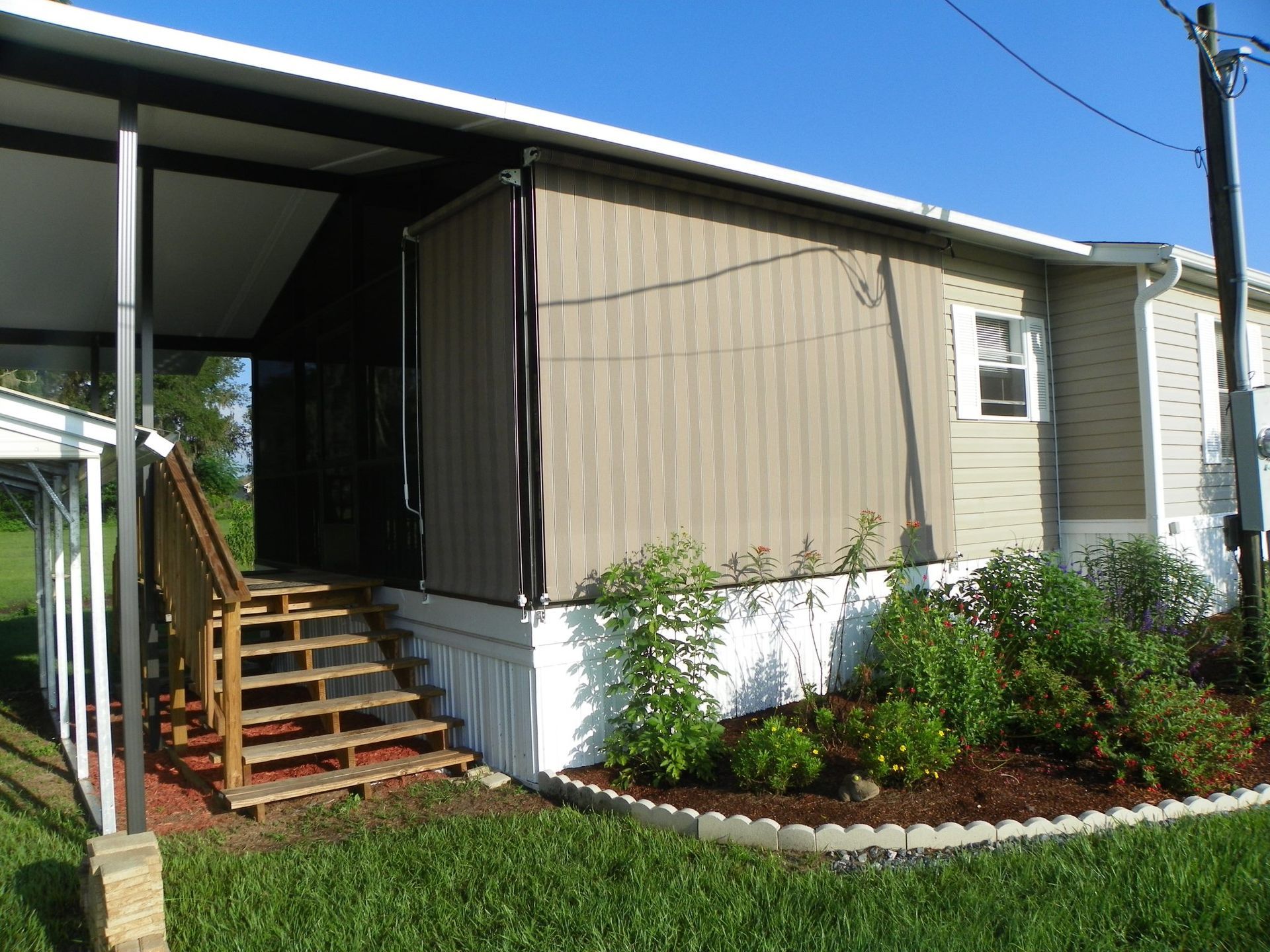 A mobile home with a beige privacy screen, a wooden staircase, and a landscaped garden bed with mulch along the base.