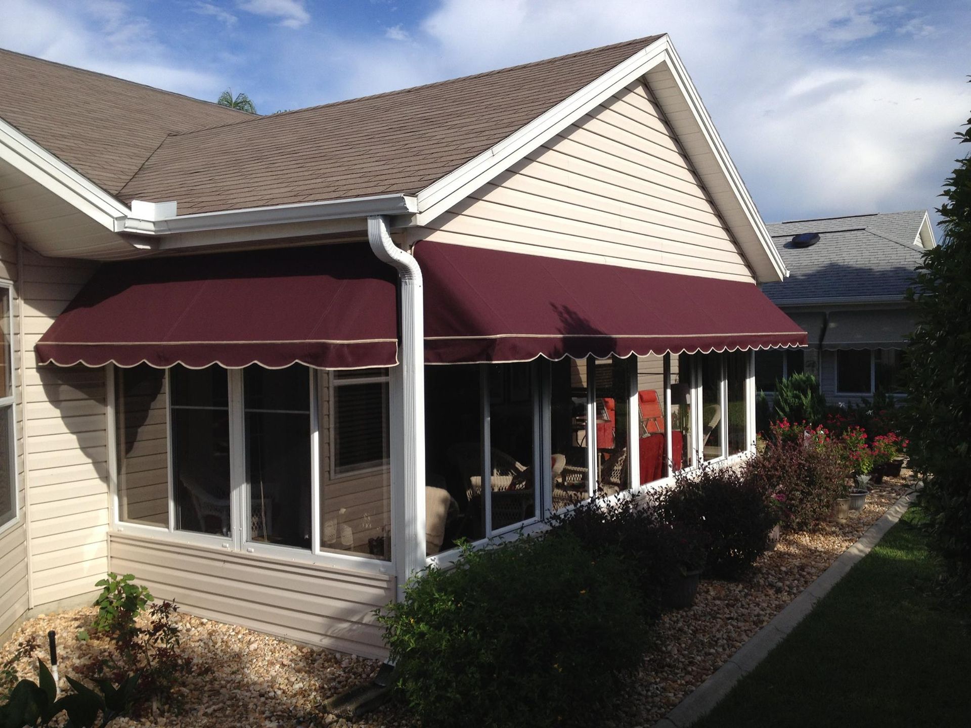 A maroon fabric awning extends over the white, windowed exterior wall of a house with tan siding and a gravel garden.