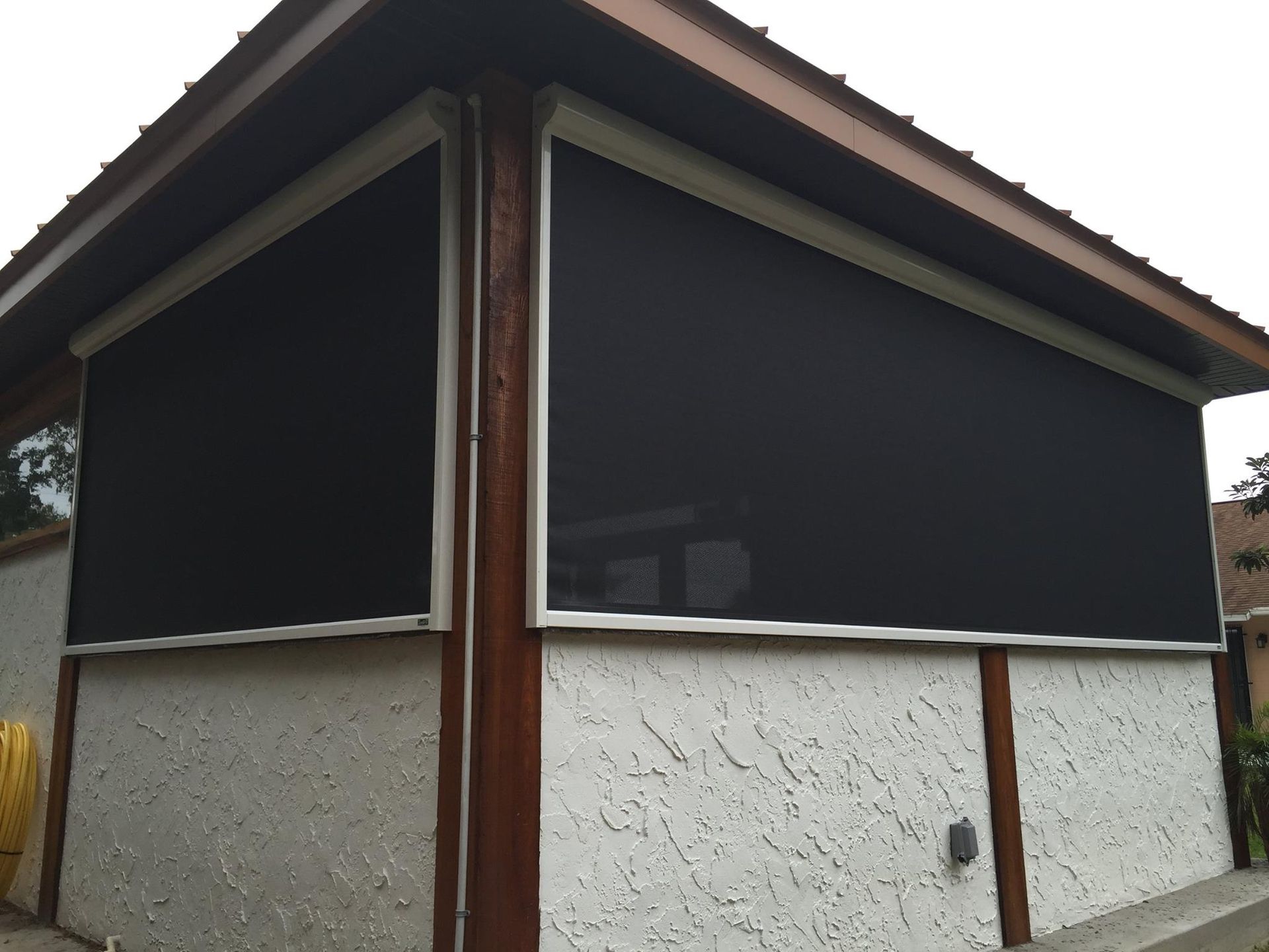 Two black solar shade screens mounted on the exterior of a home with white stucco walls and dark brown wooden trim.