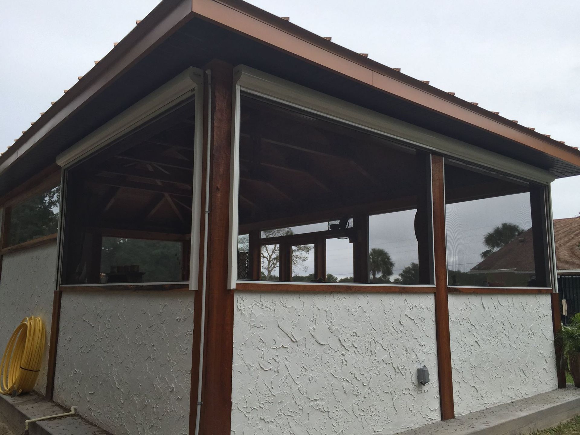 A wooden pavilion with a white textured lower wall and dark, retractable roll-down mesh screens on its open sides.