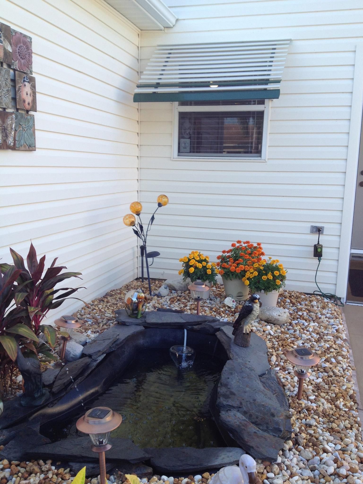 A backyard garden scene featuring a small stone pond, potted orange flowers, and solar lights against a white house wall.