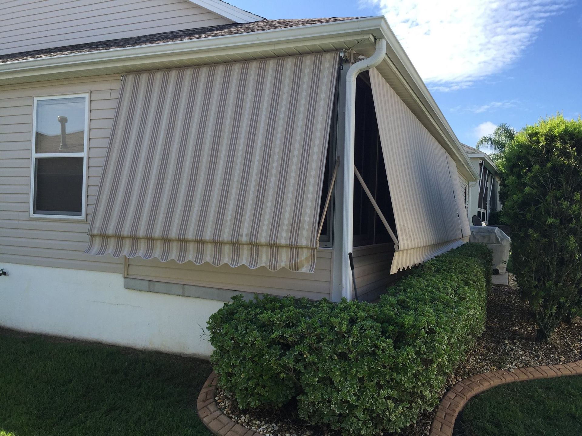 A house exterior with two beige striped awnings partially shading a screened-in porch, next to a green hedge.