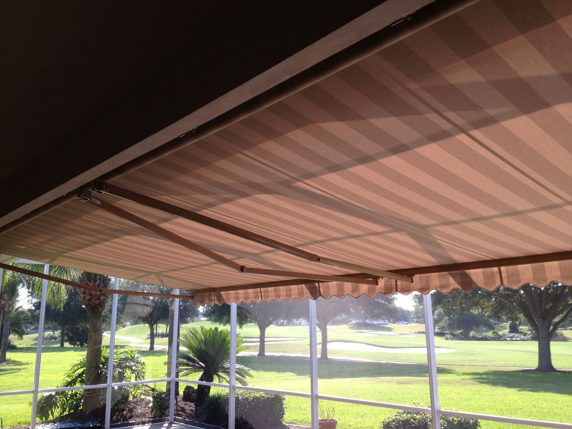 A striped tan and brown retractable awning extends over a patio, overlooking a green golf course landscape.