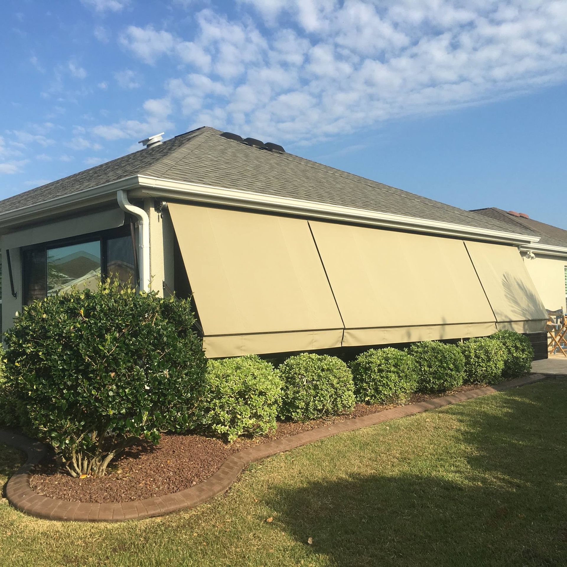 A beige retractable awning covers the side of a house behind a line of manicured green shrubs and a brown garden border.