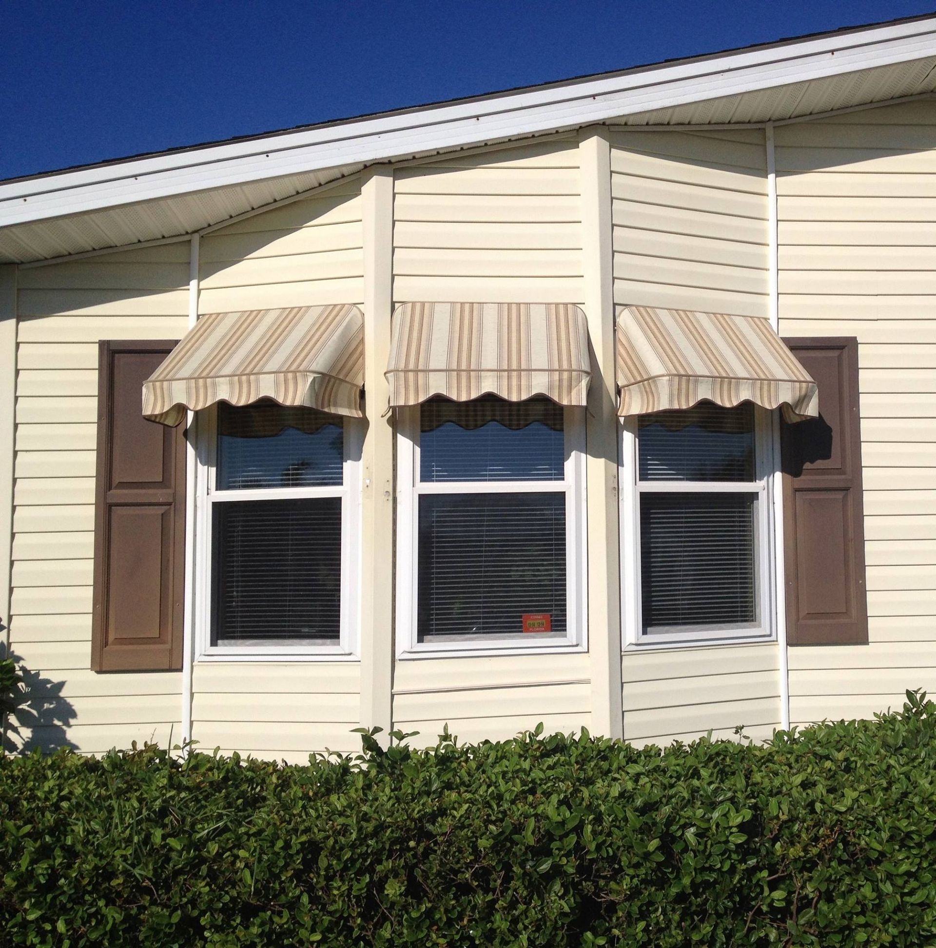 A light-yellow house exterior with three windows featuring striped fabric awnings and brown shutters, fronted by a hedge.