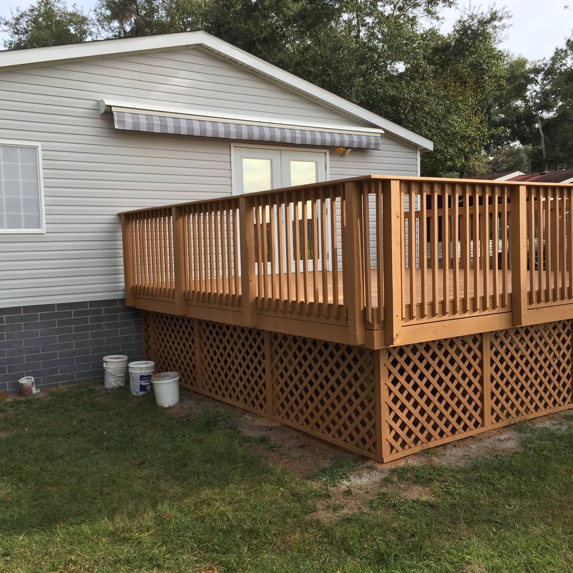 A light-colored house with a large wooden deck featuring lattice skirting, set against a grassy yard with buckets nearby.
